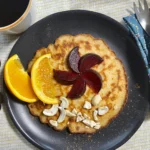 Glutenfree pancakes on a black plate decorated with plums, oranges and cashew nuts.