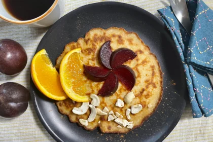 Glutenfree pancakes on a black plate decorated with plums, oranges and cashew nuts.