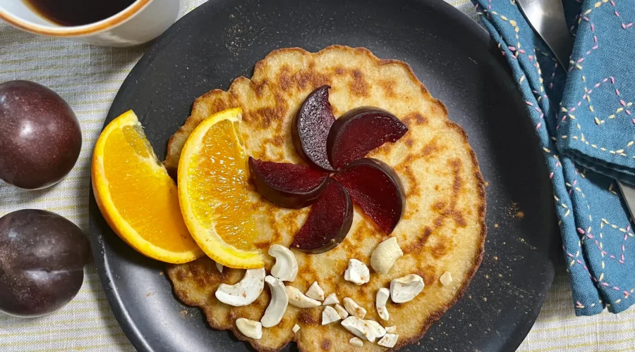 Glutenfree pancakes on a black plate decorated with plums, oranges and cashew nuts.